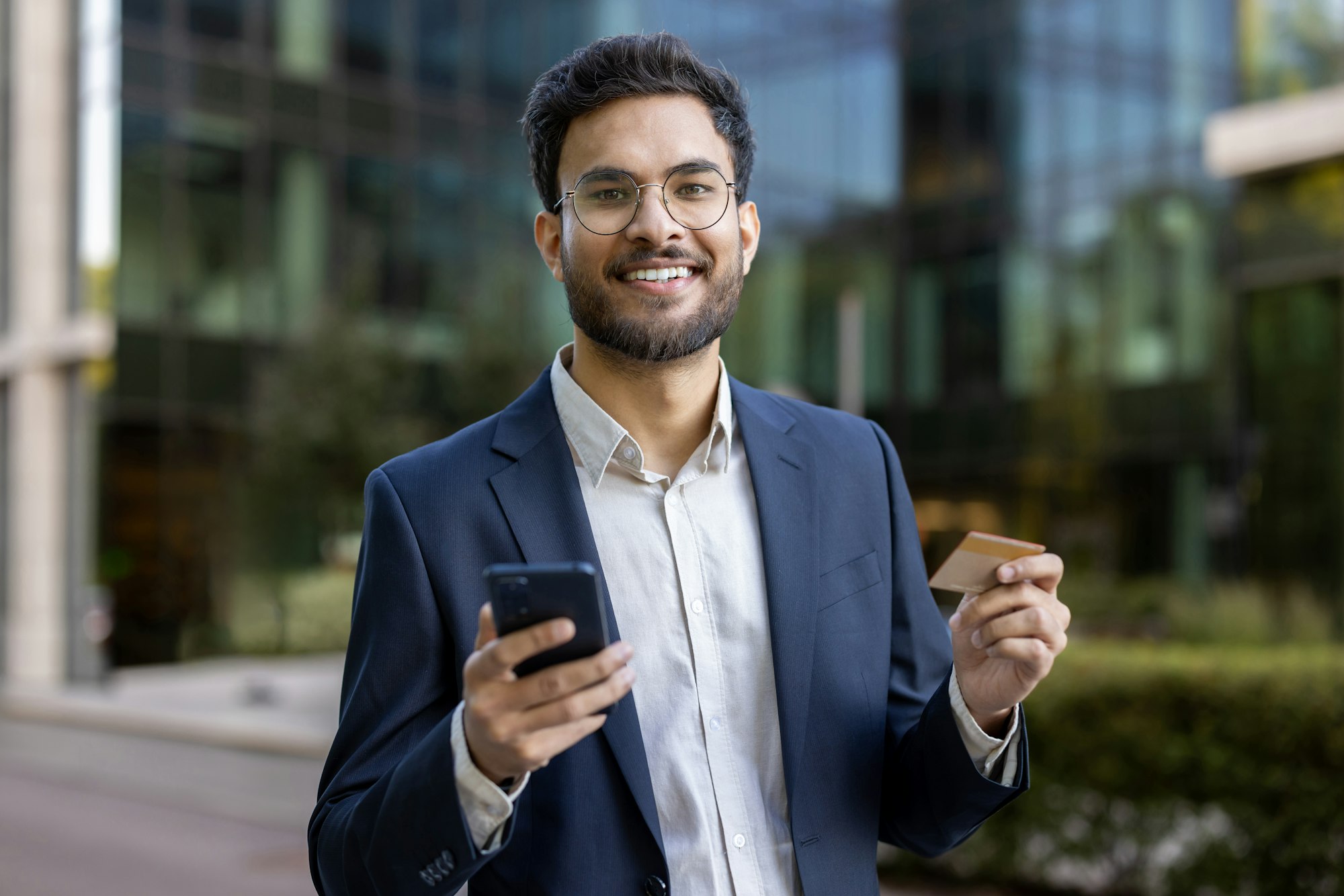 Hispanic businessman holding smartphone and credit card outdoors in modern urban setting